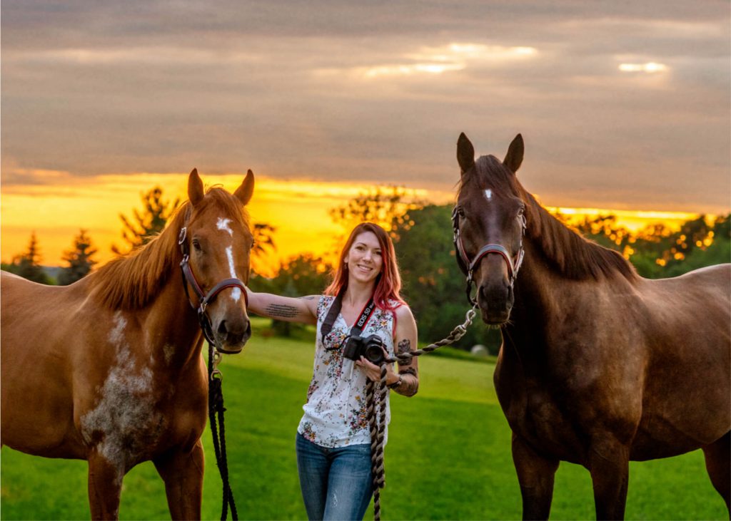 woman holding camera standing next to two horses during a sunset.